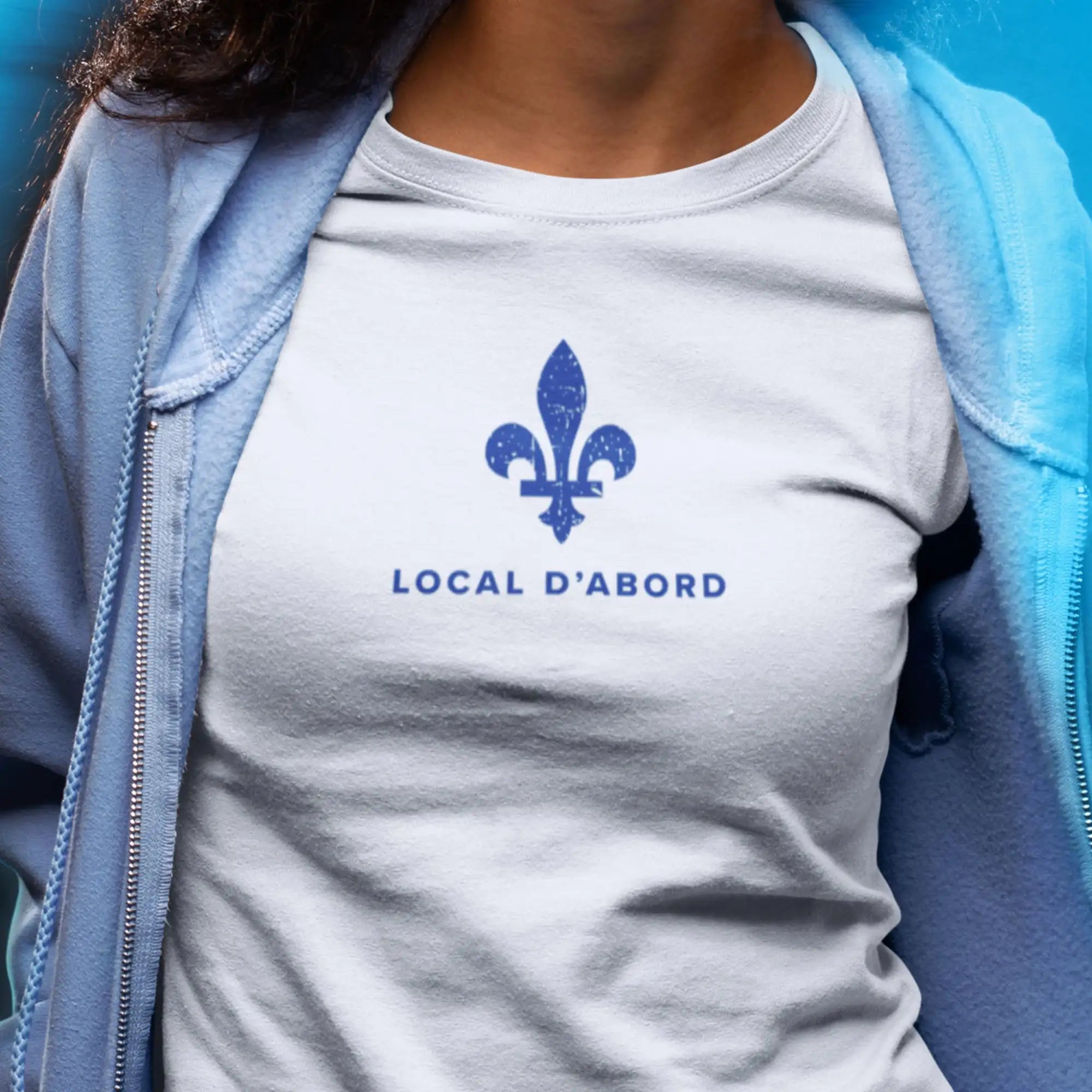 T-shirt en coton blanc ornĂ© d'un emblĂšme bleu dĂ©lavĂ© de fleur de lys et du texte « local dâabord » imprimĂ© en dessous.