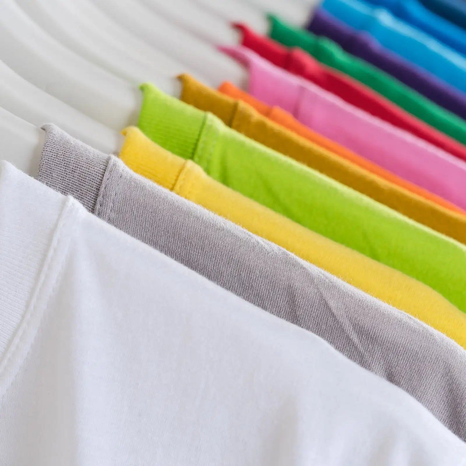 Une rangée de t-shirts colorés, pliés soigneusement, dans des teintes vives incluant le blanc, le gris, le jaune, le vert, l'orange, le rose, le rouge, le bleu et le violet.