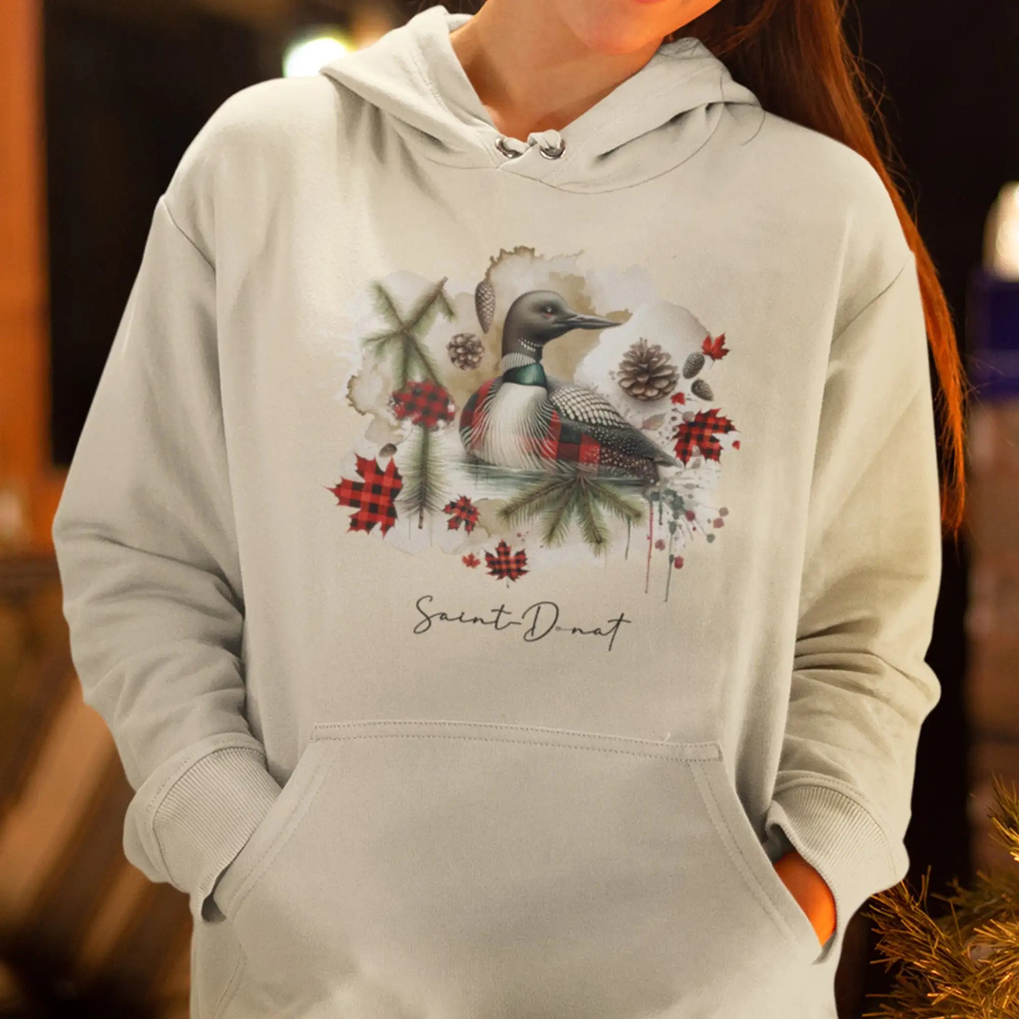Un sweat à capuche doux de couleur crème cassé orné d'un graphique détaillé d'un huard entouré de branches de pin, de feuilles à carreaux rouges et de pommes de pin, avec le texte « saint-dnat » imprimé en dessous.