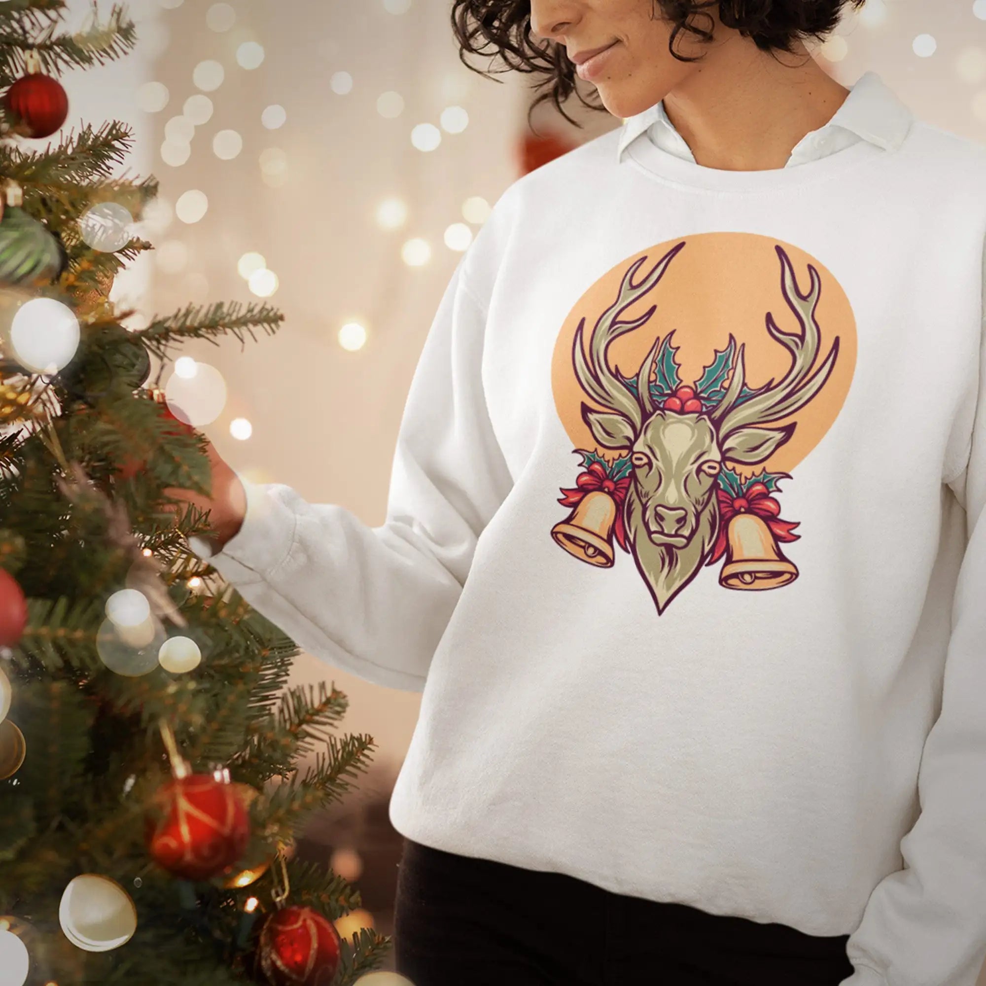 Sweatshirt à col rond blanc orné d'un graphique coloré représentant la tête d'un cerf avec des bois, décorée de houx et de deux cloches sur un fond circulaire orange.