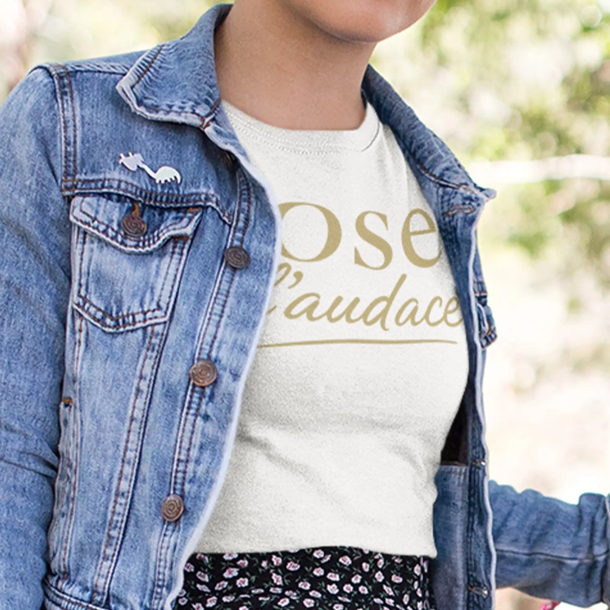 T-shirt blanc avec lettrage doré indiquant « ose l'audace » et une petite broderie de dinosaure blanc sur la poche poitrine de la veste en denim.