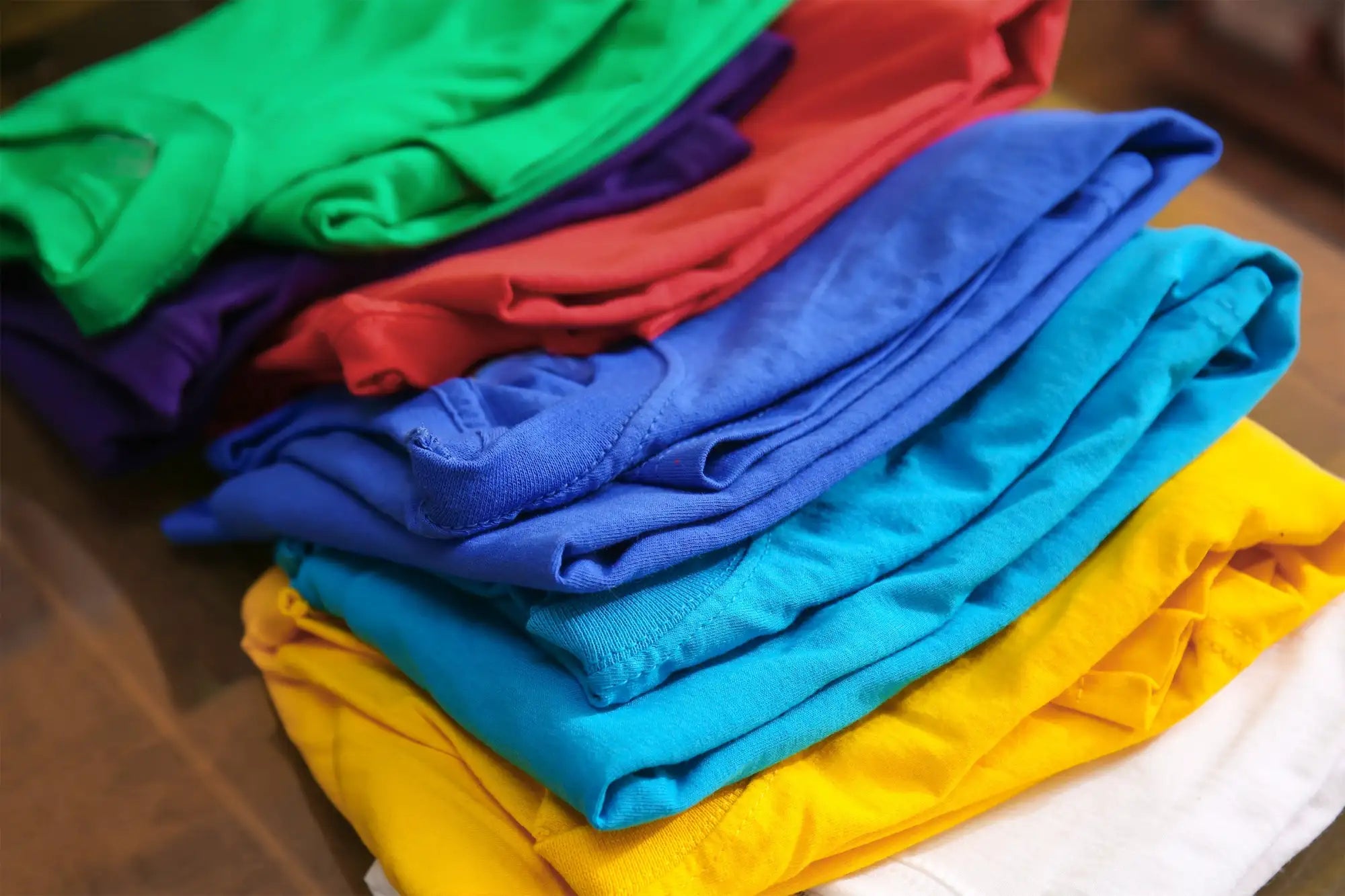 Tas de t-shirts colorés soigneusement pliés en vert vibrant, rouge, bleu, turquoise, jaune et blanc.